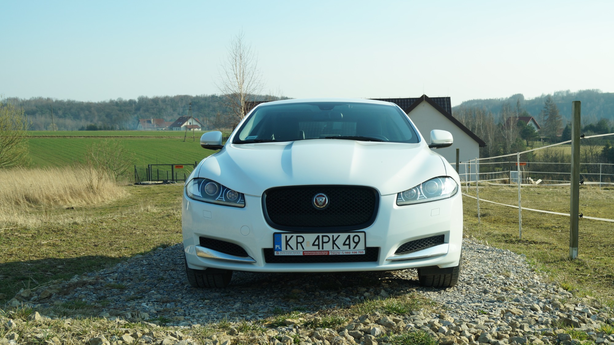 Biały samochód Jaguar XF z tablicą rejestracyjną KR 4PK49, zaparkowany na żwirowej nawierzchni przed wiejskim krajobrazem z budynkami i polami.
