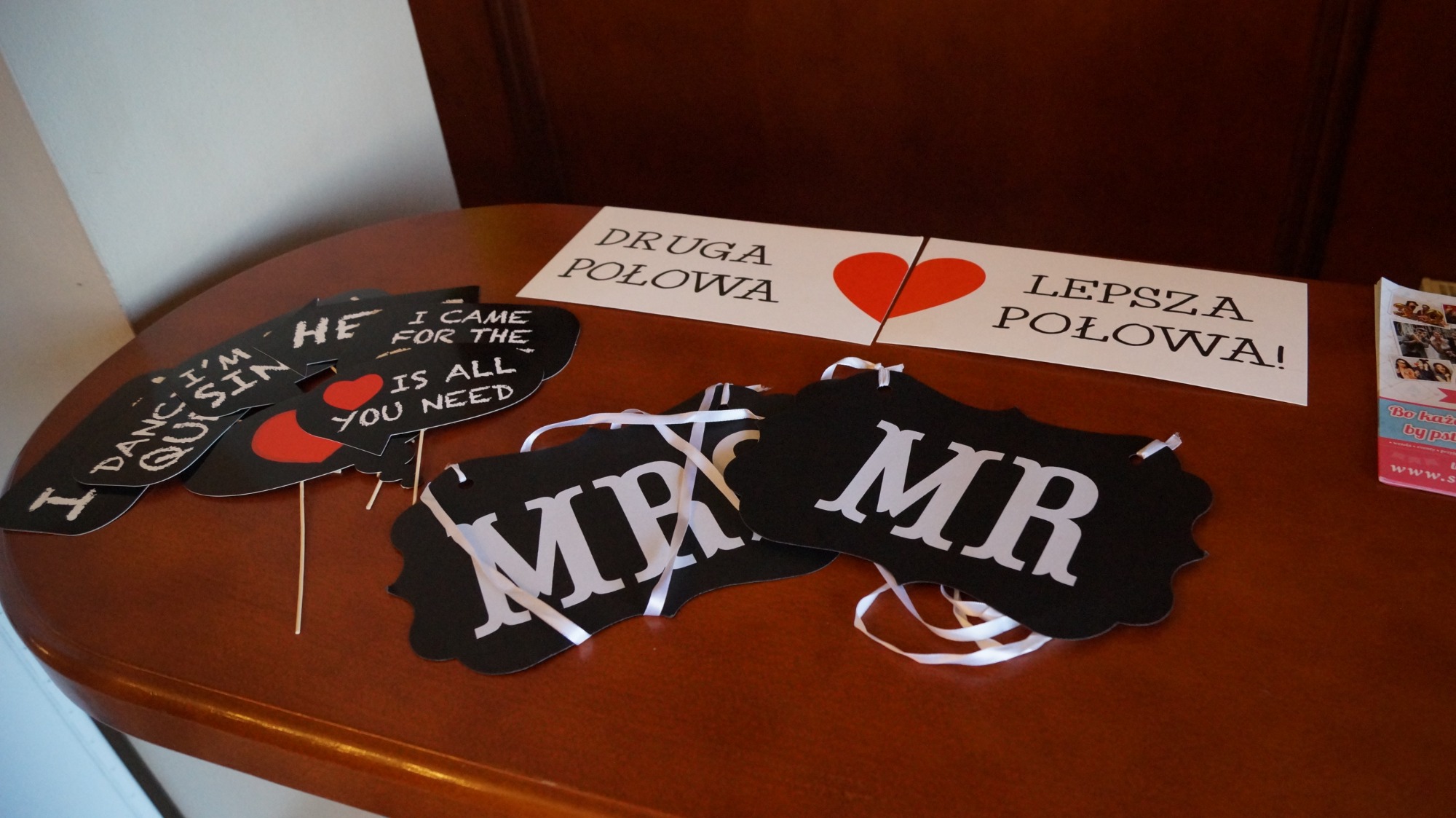 Rekwizyty do fotobudki na brązowym stole: czarne tabliczki z napisami 'MR' na białych wstążkach, 'I DANC. I'M QUISIN', 'HE', 'I CAME FOR THE IS ALL YOU NEED' z czerwonym sercem, oraz kartki...