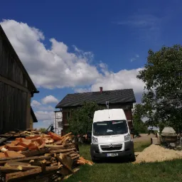 Biały samochód dostawczy na tle domu z nowym dachem, sterta drewnianych elementów konstrukcyjnych obok, widoczne rusztowania i drabina na dachu.