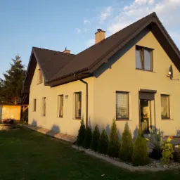 Ocieplony dom jednorodzinny z brązowym dachem i jasną elewacją, widok z ogrodu w ciepłym świetle popołudniowym, z widoczną anteną satelitarną.