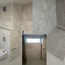 Nowoczesna łazienka z szarymi płytkami w różnych odcieniach i wzorach, widoczny odpływ liniowy, podwieszana toaleta z przyciskiem spłukującym i wentylator wyciągowy.