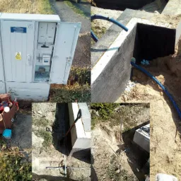 Otwarta szafa elektryczna z widocznymi zabezpieczeniami, obok narzędzia elektryczne i fundament budynku z niebieskimi rurami ochronnymi na kable w ziemi.