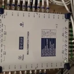 Multiswitch TT-924 z licznymi kablami koncentrycznymi podłączonymi do wejść i wyjść, widoczne oznaczenia portów i tabela częstotliwości, instalacja w szarej szafie rozdzielczej.