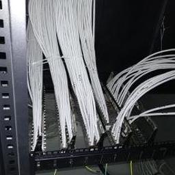 ASEL INSTALACJE - Rząd białych kabli sieciowych uporządkowanych w szafie rack, widok z dołu, ciemne tło, metalowa konstrukcja szafy po lewej stronie.