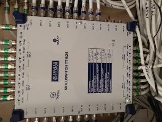Multiswitch TT-924 z licznymi kablami koncentrycznymi podłączonymi do wejść i wyjść, widoczne oznaczenia portów i tabela częstotliwości, instalacja w szarej szafie rozdzielczej.