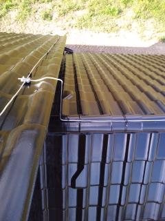 Fragment dachu z zamontowanym systemem odgromowym: przewód odprowadzający przymocowany do rynny i uchwyt na dachówce ceramicznej.