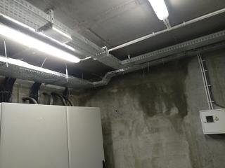 Szare, betonowe pomieszczenie techniczne z widocznymi korytami kablowymi pod sufitem, białym panelem elektrycznym na ścianie i fragmentem szafy sterowniczej. Widoczne ślady wilgoci na ścianie.