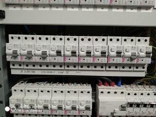 Rozdzielnica elektryczna z widocznymi bezpiecznikami automatycznymi i przewodami. Górny rząd bezpieczników z opisami, dolny rząd częściowo zasłonięty. Widoczne fragmenty obudowy rozdzielnicy.