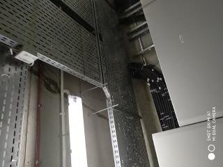 Metalowe koryta kablowe zamocowane pod sufitem w surowym wnętrzu budynku, widoczne przewody i elementy konstrukcyjne, oświetlenie fluorescencyjne.