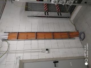 Pomarańczowe przewody ułożone równolegle w metalowej korycie kablowej przytwierdzonej do ściany z białych bloczków. Widoczny fragment sufitu z konstrukcją podwieszaną i inne kable. Przy ścianie...