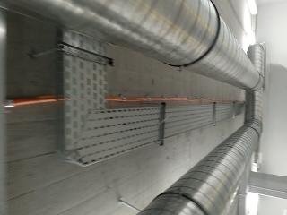 Metalowe koryta kablowe z perforacją, zamontowane na betonowej ścianie, widoczne przewody wewnątrz, obok rury wentylacyjne ze stali nierdzewnej, ujęcie z dołu.