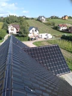 Czarny, błyszczący dach z widoczną instalacją odgromową na tle zielonego wzgórza z zabudową jednorodzinną w słoneczny dzień.