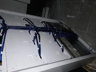 Rozdzielnica elektryczna z widocznymi przewodami w niebieskiej peszli, białe panele z naklejkami identyfikacyjnymi, ujęcie z góry pod kątem.