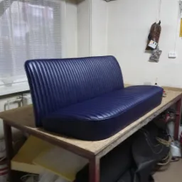 Nowe, granatowe obicie kanapy samochodowej z pionowymi przeszyciami, leżące na warsztatowym stole, z widocznym kompresorem i narzędziami w tle.