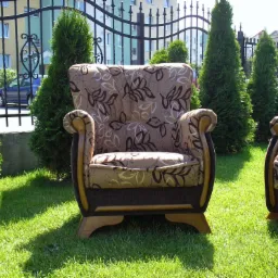 Odnowiony fotel z brązową tapicerką w kwiaty, ustawiony na trawniku przed metalowym ogrodzeniem i tujami.