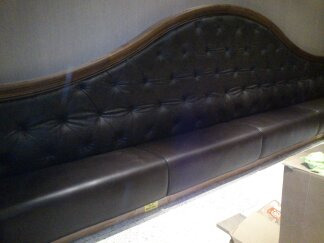 Długa, ciemnobrązowa sofa z pikowanym oparciem i drewnianą ramą, widoczna z boku, z elementami kartonu i szmatki w tle po prawej stronie.