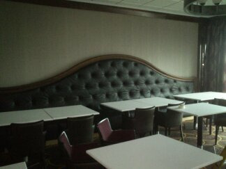 Tapicerowana, pikowana ława w restauracji z ciemnobrązową ramą i beżową ścianą w tle, uzupełniona białymi stolikami i krzesłami.