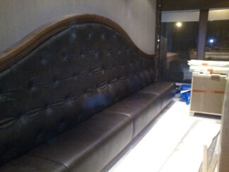 Długa, ciemna kanapa z pikowanym oparciem i drewnianą ramą, ustawiona w pomieszczeniu z widocznym oknem i kartonami w tle.