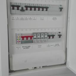 Remonty Wykończenia - Otwarta, biała skrzynka elektryczna z bezpiecznikami i wyłącznikami oznaczonymi dla różnych pomieszczeń, w tym salonu, łazienki i kuchni, z włącznikiem głównym i kontrolką faz. Część wyłączników...