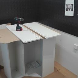 Remonty Wykończenia - Montaż szafki łazienkowej w trakcie, widoczna wiertarka akumulatorowa na blacie, instrukcja wewnątrz szafki, czarne płytki na ścianie w tle.