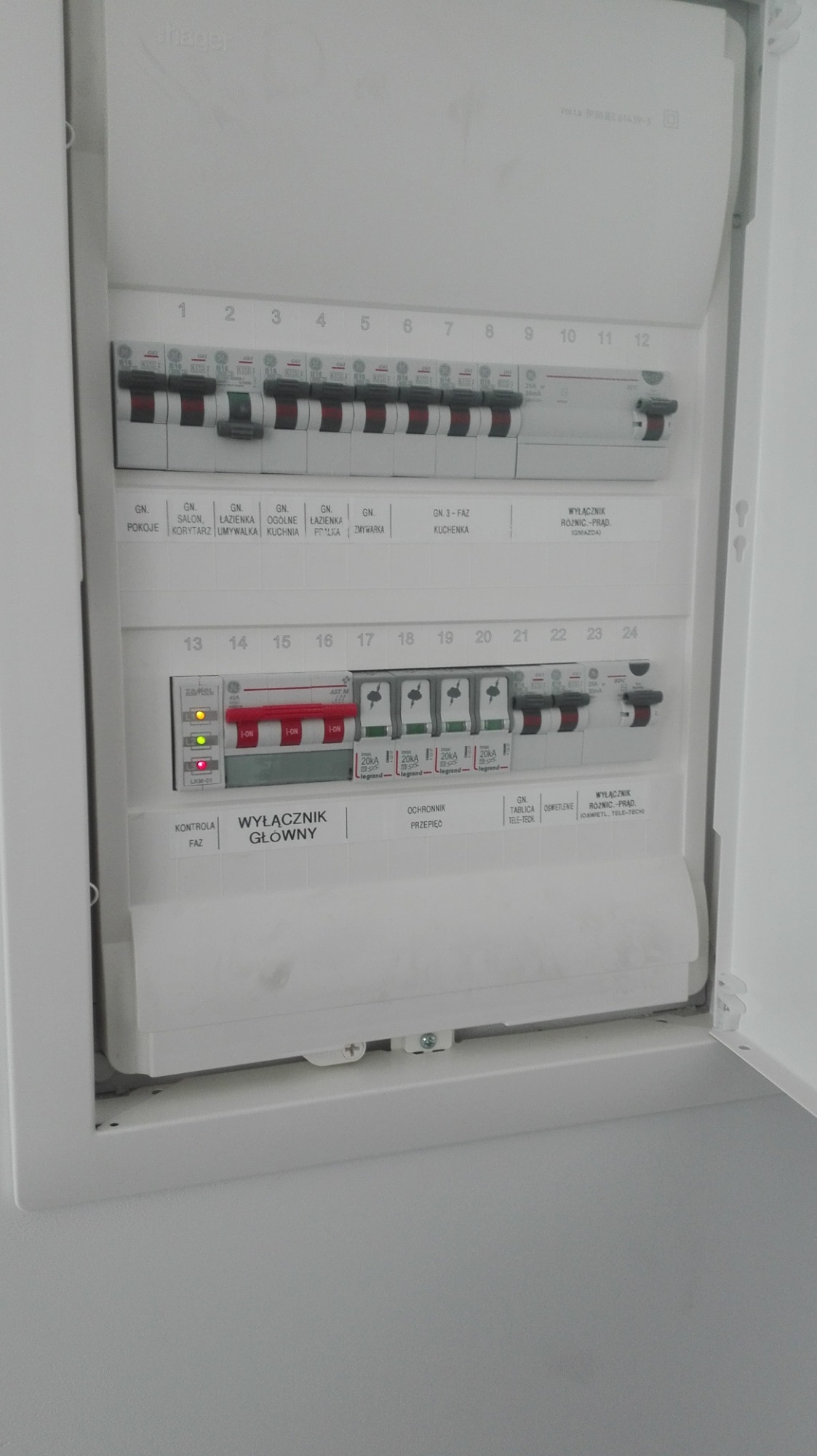 Otwarta, biała skrzynka elektryczna z bezpiecznikami i wyłącznikami oznaczonymi dla różnych pomieszczeń, w tym salonu, łazienki i kuchni, z włącznikiem głównym i kontrolką faz. Część wyłączników...