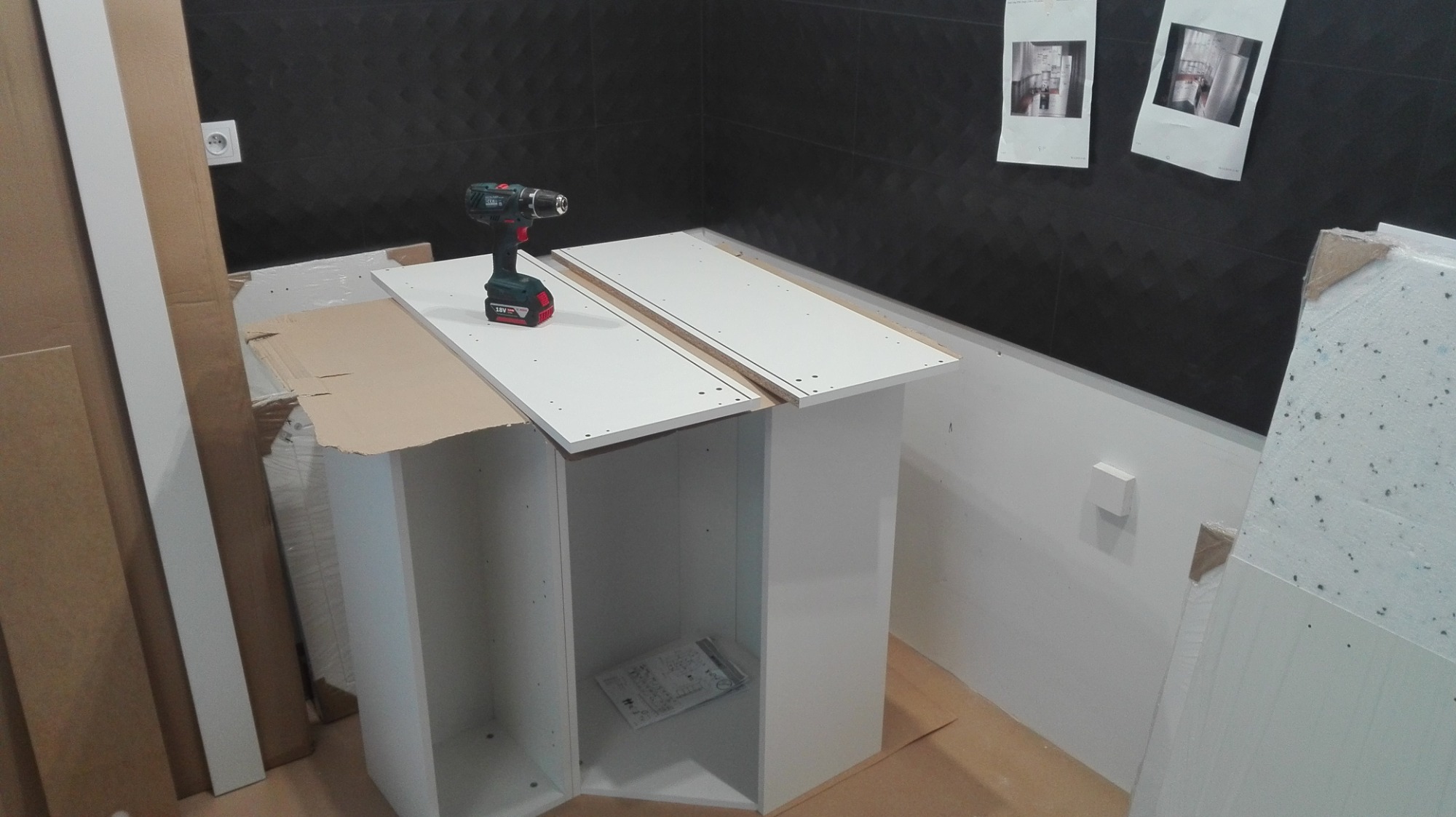Montaż szafki łazienkowej w trakcie, widoczna wiertarka akumulatorowa na blacie, instrukcja wewnątrz szafki, czarne płytki na ścianie w tle.