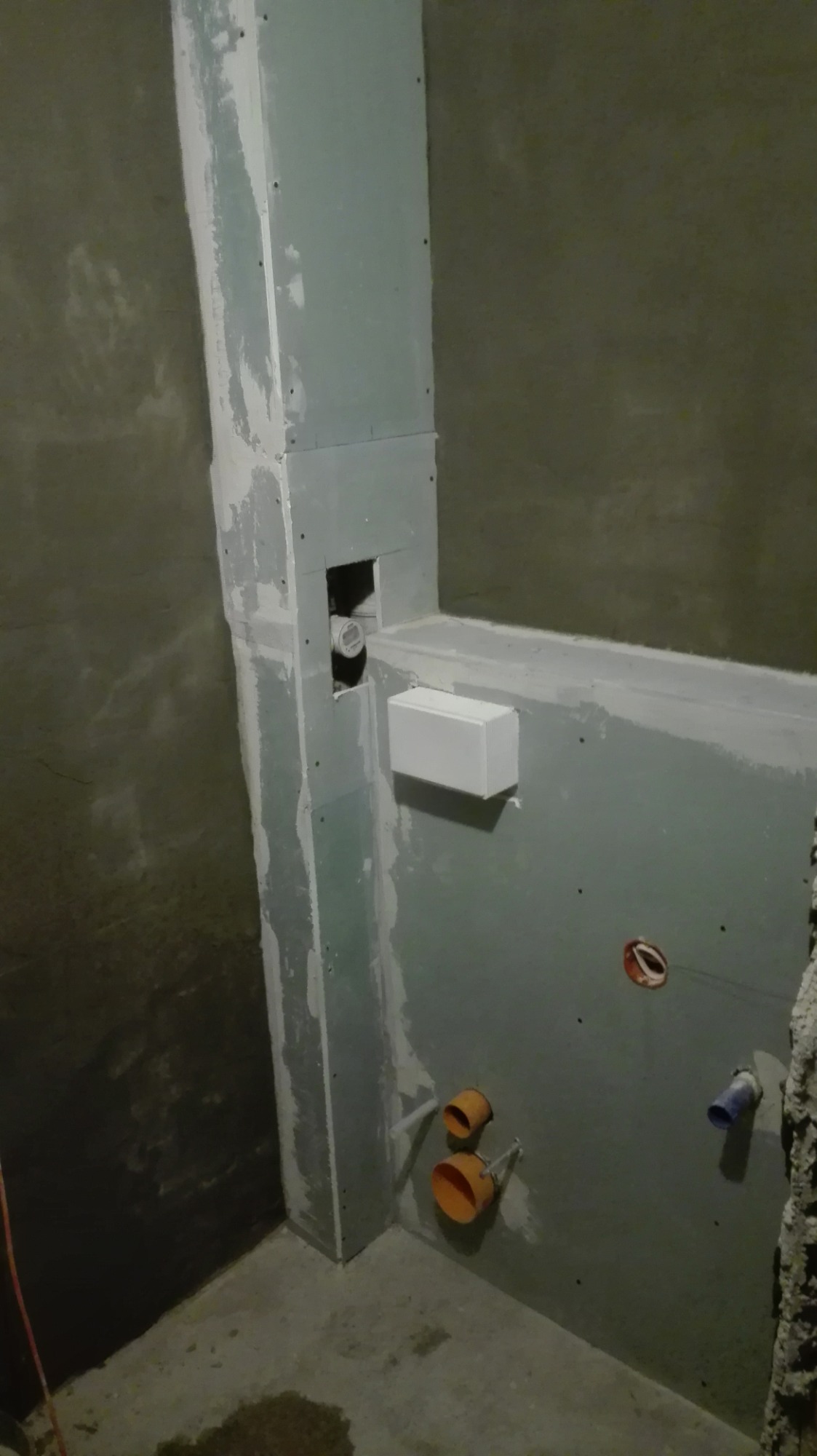 Surowy stan łazienki w trakcie prac: widoczne płyty gipsowo-kartonowe na ścianach, wystające rury kanalizacyjne w kolorze pomarańczowym i niebieskim, oraz puszka elektryczna bez osprzętu.