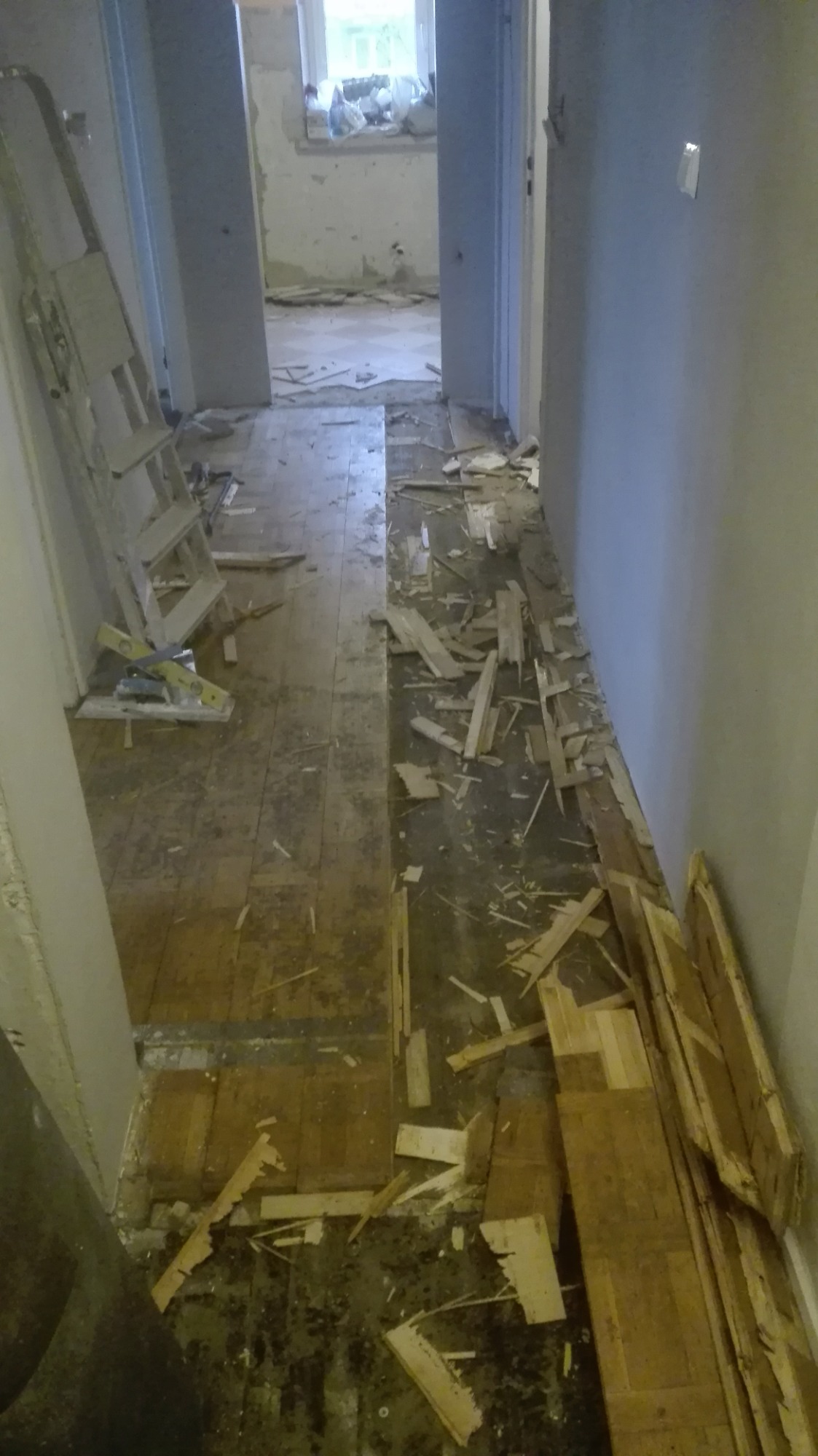 Demontaż starego parkietu w korytarzu, widoczna drabina i poziomica, podłoga częściowo zerwana, gruz i deski na podłodze.