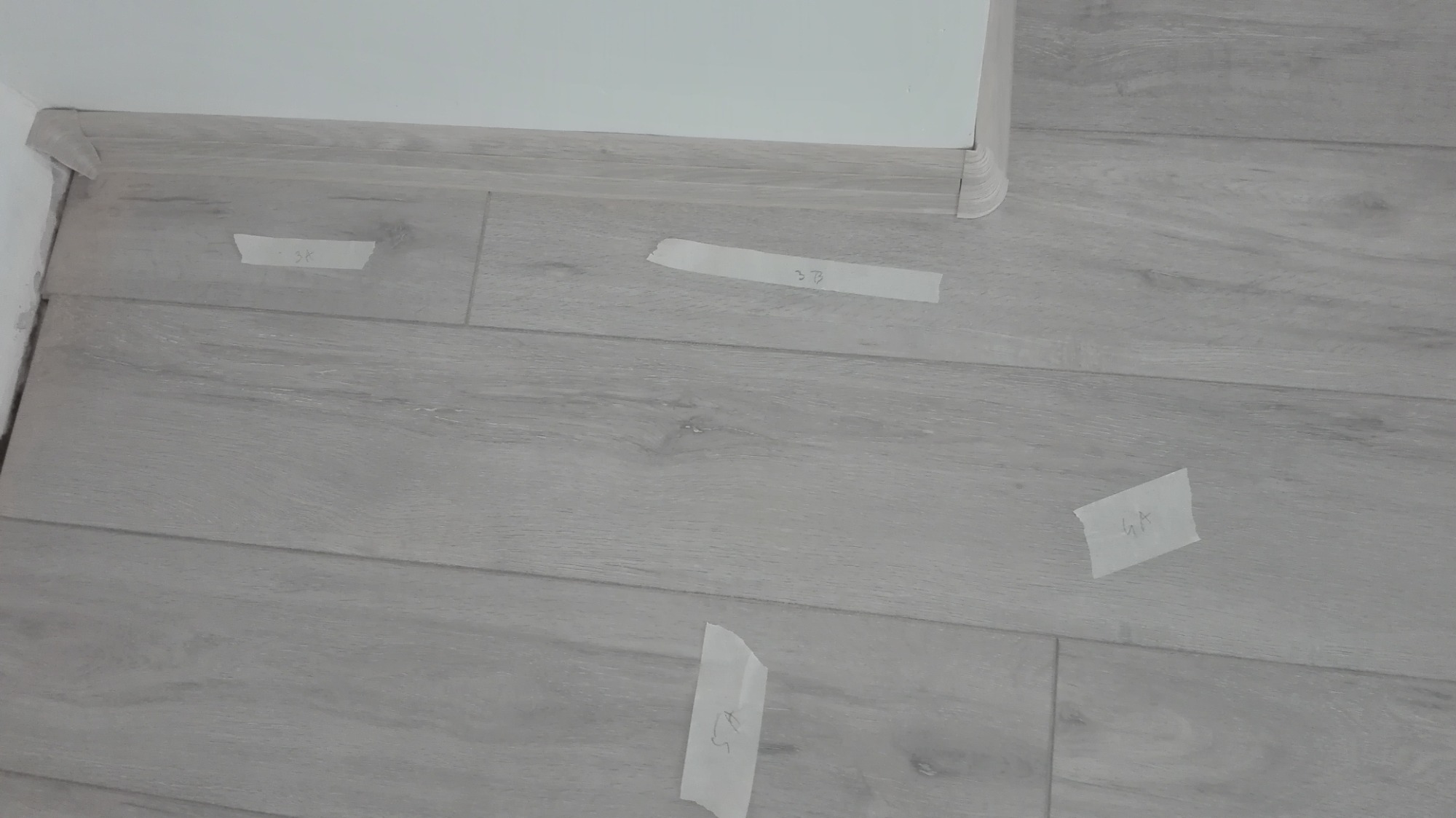 Jasne panele podłogowe z przyklejonymi taśmami z numerami, widoczne listwy przypodłogowe.