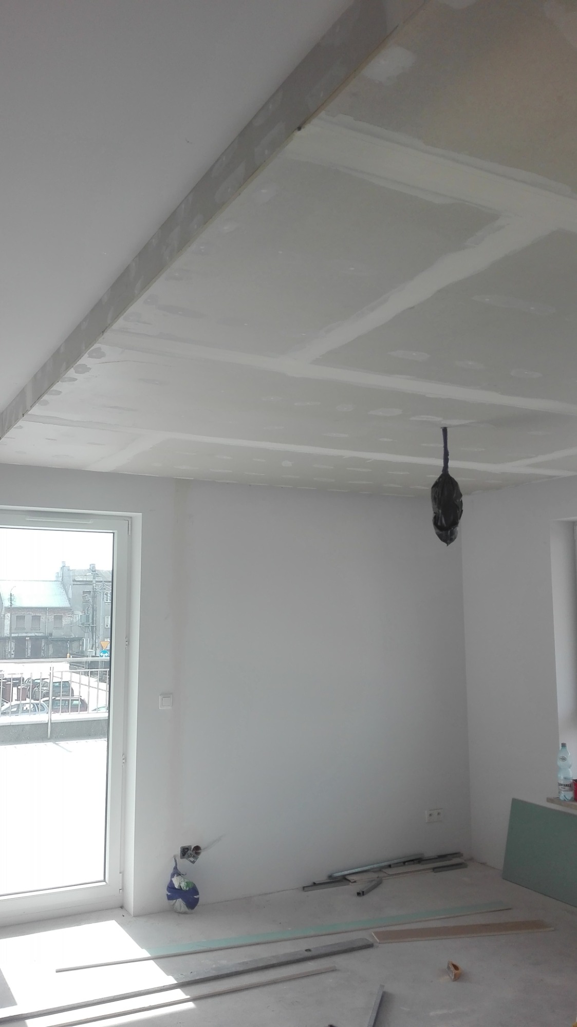 Wnętrze w trakcie prac: sufit z płyt gipsowo-kartonowych, widoczne łączenia wymagające szpachlowania, wystające kable elektryczne zabezpieczone workiem, profile metalowe na podłodze, okno balkonowe...