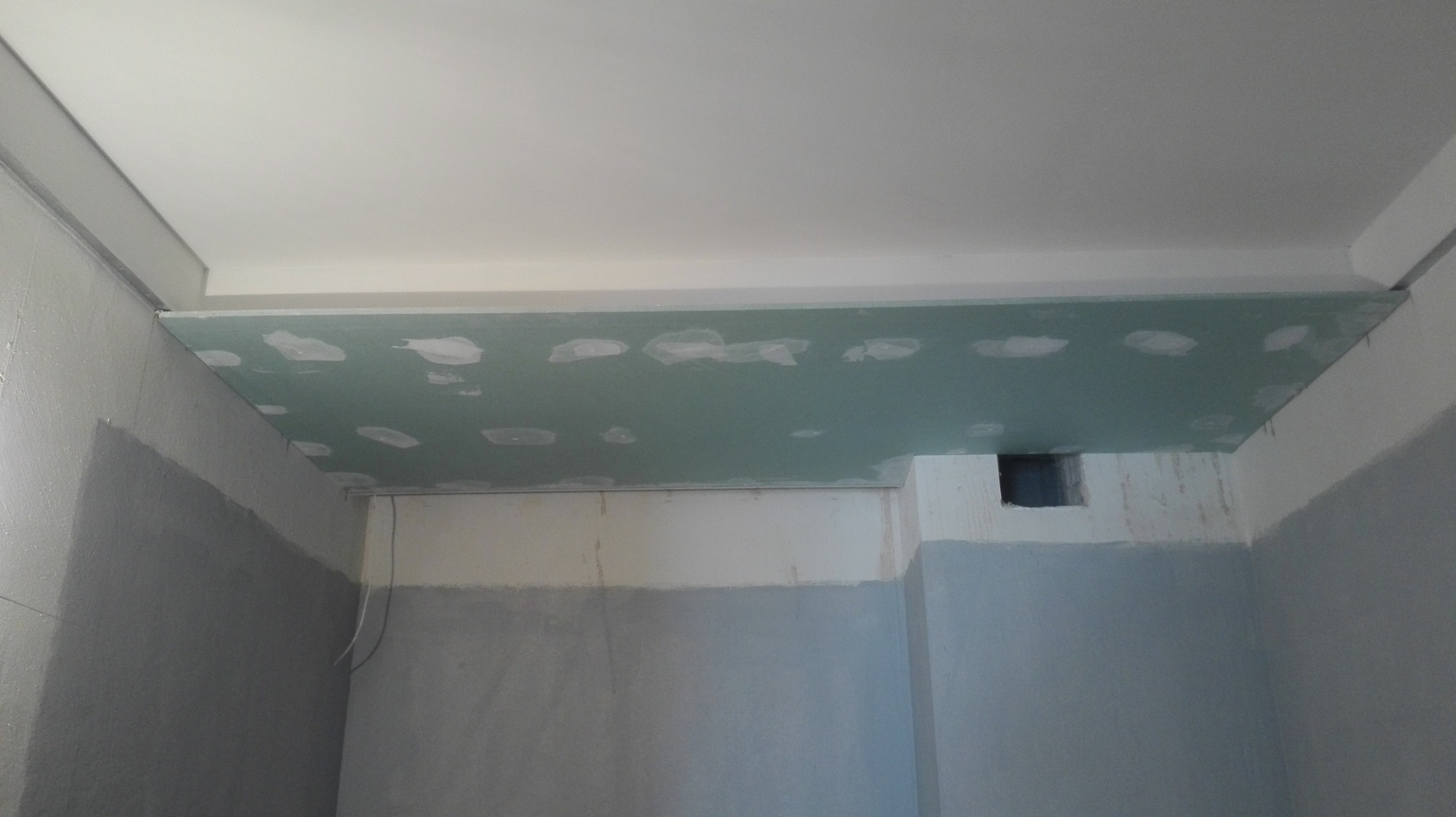 Sufit podwieszany z płyty gipsowo-kartonowej w trakcie montażu, widoczne łączenia szpachlowane, surowe ściany w odcieniach szarości i bieli, fragment instalacji wentylacyjnej.