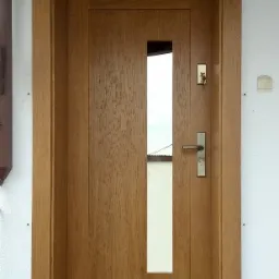 Dębowa stolarka z wąskim przeszkleniem i metalową klamką, zamontowana w białej ścianie zewnętrznej budynku.