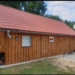 Drewniany budynek z czerwoną dachówką, oświetlony zewnętrznymi lampami, z widoczną anteną satelitarną i elementami wykończenia.