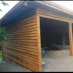 Drewniana wiata z deskami na ścianach, widoczne dwie taczki wewnątrz, betonowy fundament i fragment trawnika.