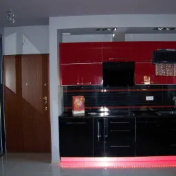 Nowoczesna kuchnia z czerwonymi szafkami górnymi i czarnymi dolnymi, podświetlana czerwonymi diodami LED, z dekoracyjnym żyrandolem kryształowym i przesuwnymi drzwiami szafy z lustrem w tle.