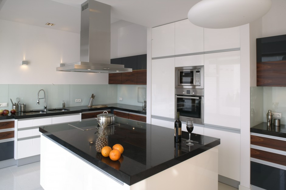 Nowoczesna kuchnia z czarnym blatem wyspowym, białymi szafkami, stalowym okapem i owocami oraz winem na blacie.
