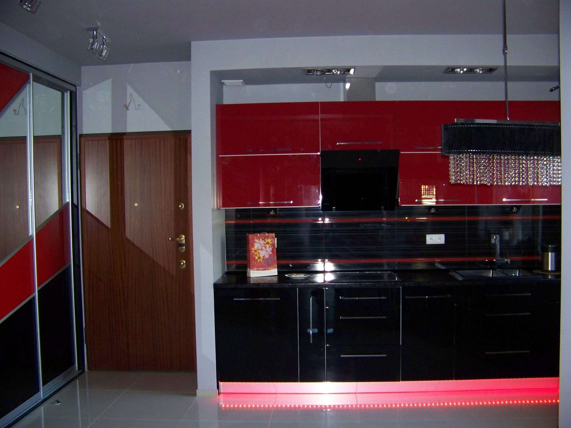 Nowoczesna kuchnia z czerwonymi szafkami górnymi i czarnymi dolnymi, podświetlana czerwonymi diodami LED, z dekoracyjnym żyrandolem kryształowym i przesuwnymi drzwiami szafy z lustrem w tle.