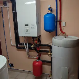 Zainstalowany, wiszący na ścianie kocioł grzewczy Panasonic z podłączonymi instalacjami: zbiornikiem wyr&oacute;wnawczym w kolorze niebieskim, zasobnikiem ciepłej wody i instalacją hydrauliczną w kolorach...