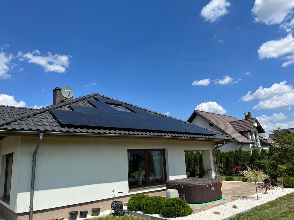 Czarny panel słoneczny na dachu domu jednorodzinnego z brązową dachówką, widok z ogrodu w słoneczny dzień, w tle błękitne niebo i sąsiedni dom.