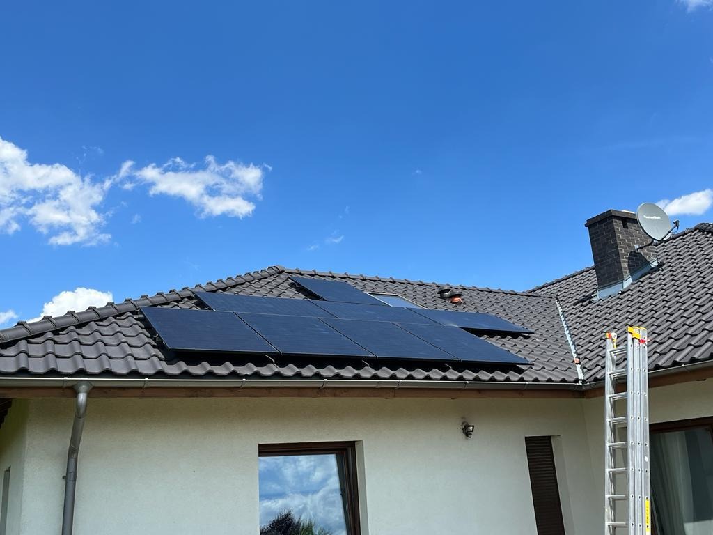 Panele słoneczne na dachu domu jednorodzinnego z brązową dachówką, widoczny komin, antena satelitarna i przystawiona drabina aluminiowa.