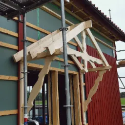 Budowa zadaszenia nad wejściem do budynku, widoczne drewniane elementy konstrukcyjne i rusztowanie.