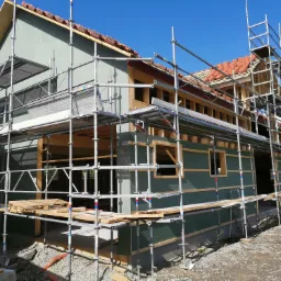 Dom w trakcie budowy z widocznym rusztowaniem i częściowo ułożoną czerwoną dachówką, ściany zewnętrzne pokryte zielonym materiałem izolacyjnym.