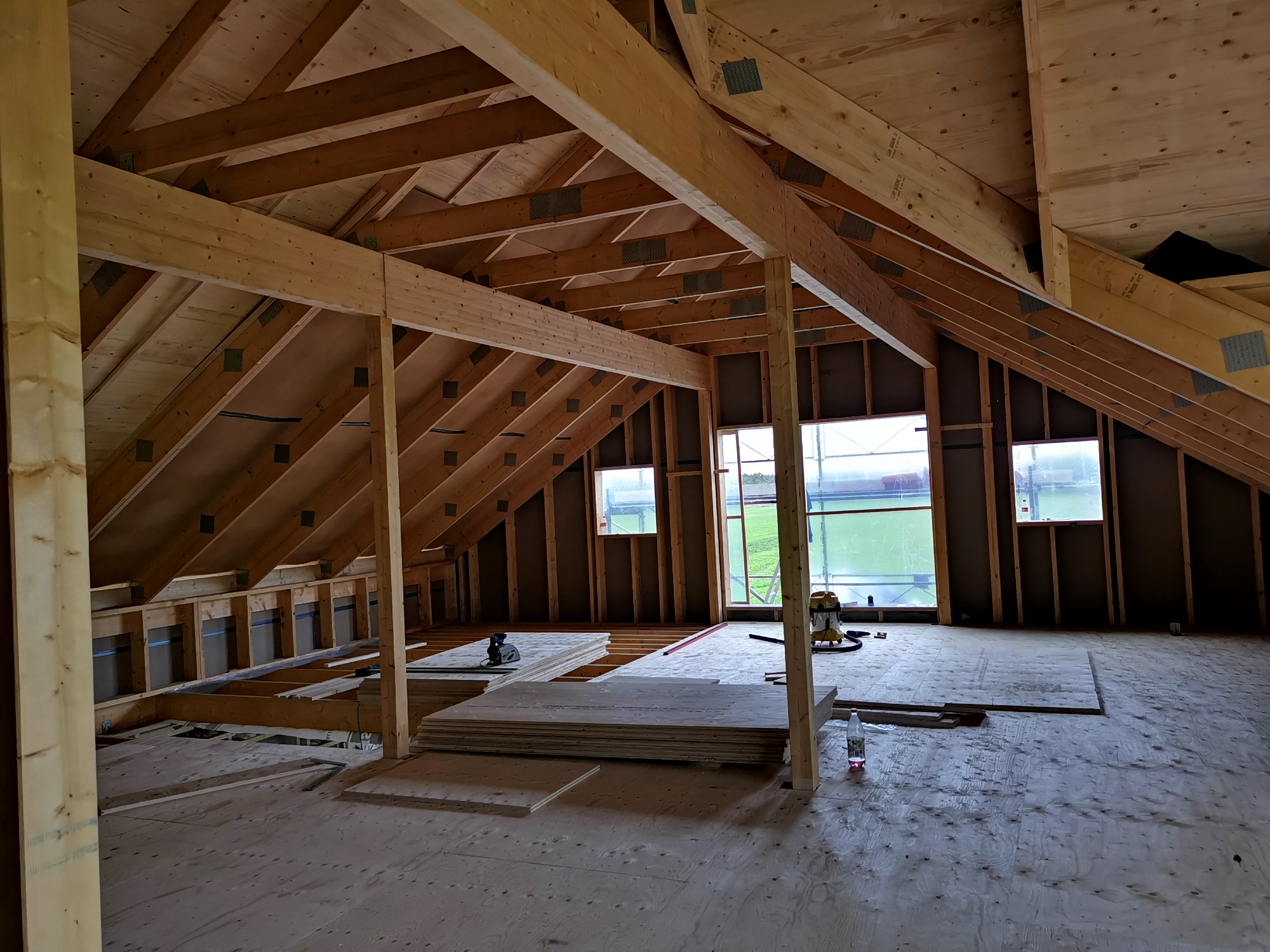 Wnętrze poddasza w trakcie budowy z widoczną drewnianą konstrukcją dachu, oknami z widokiem na zewnątrz i przygotowanymi płytami na podłodze.