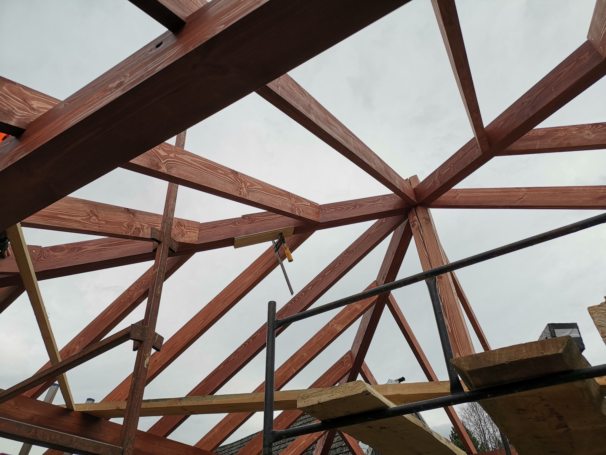 Konstrukcja dachu z belek drewnianych w kolorze brązowym, widok od dołu, elementy rusztowania w tle