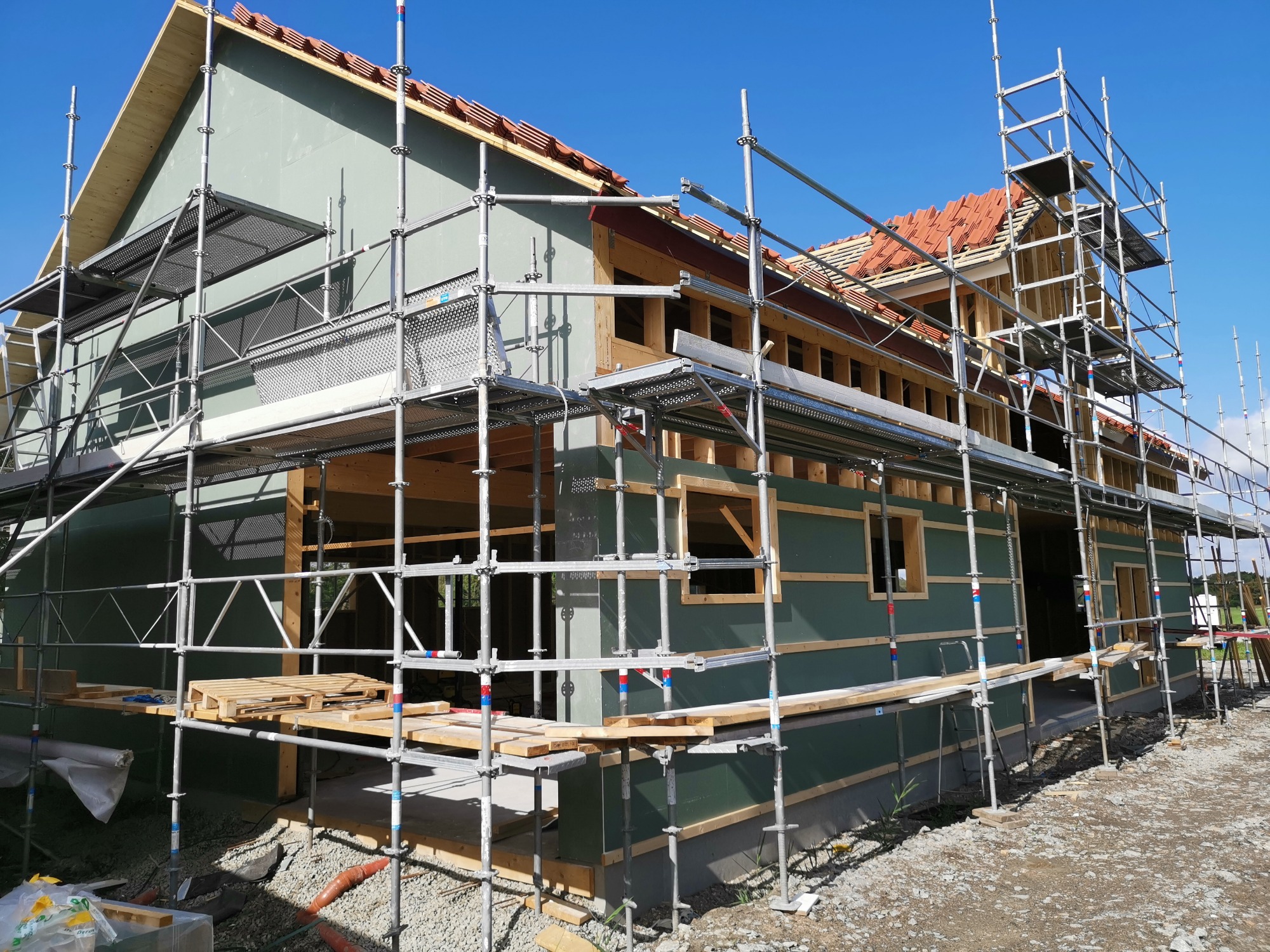 Dom w trakcie budowy z widocznym rusztowaniem i częściowo ułożoną czerwoną dachówką, ściany zewnętrzne pokryte zielonym materiałem izolacyjnym.