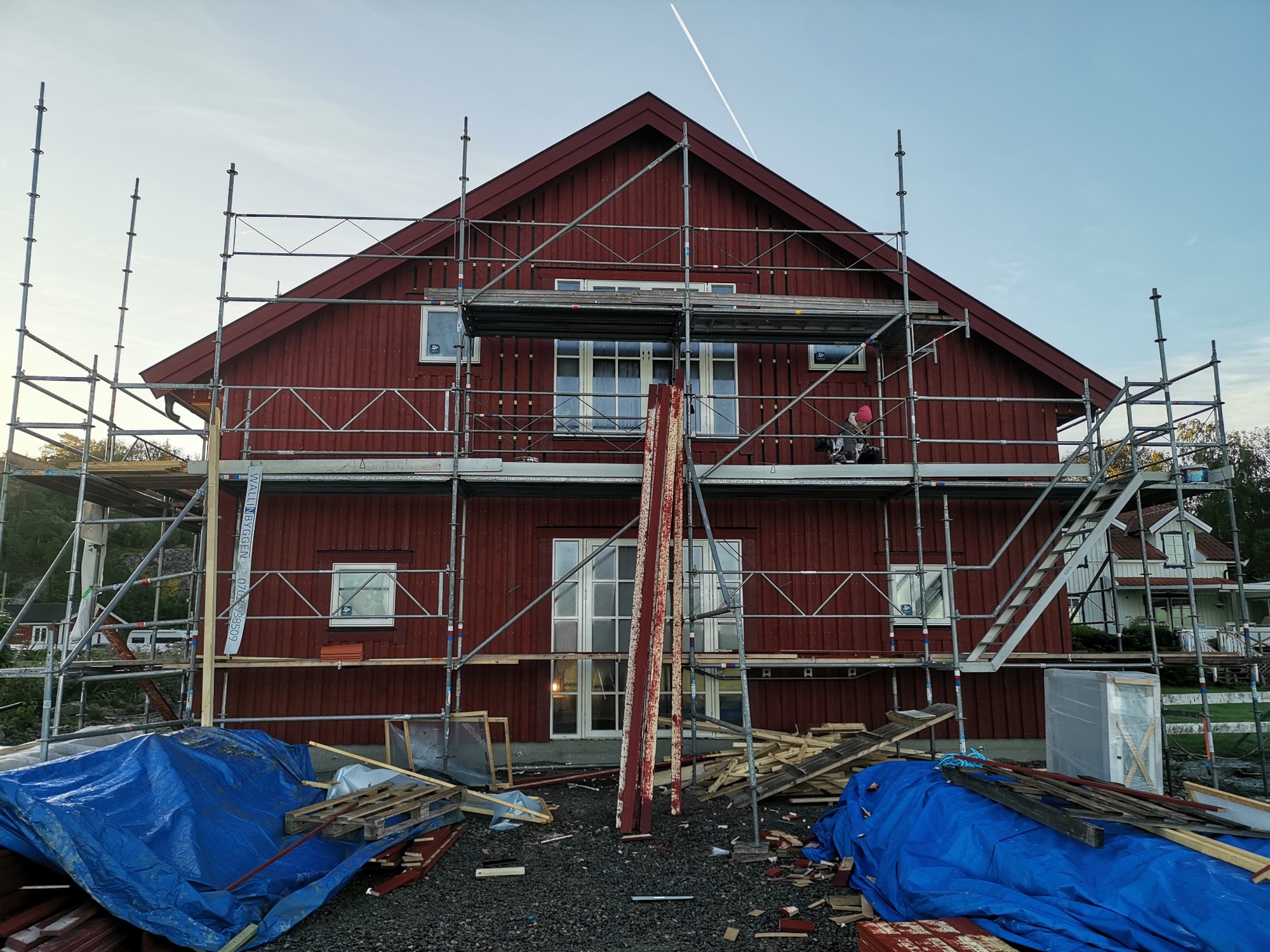 Czerwony dom w trakcie remontu dachu, otoczony rusztowaniami, z niebieskimi plandekami i gruzem budowlanym na ziemi, widoczny ślad kondensacyjny samolotu na niebie.