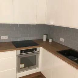 Nowoczesna kuchnia z białymi, lakierowanymi frontami, drewnianym blatem, szarą glazurą typu metro i czarnym zlewem kompozytowym.