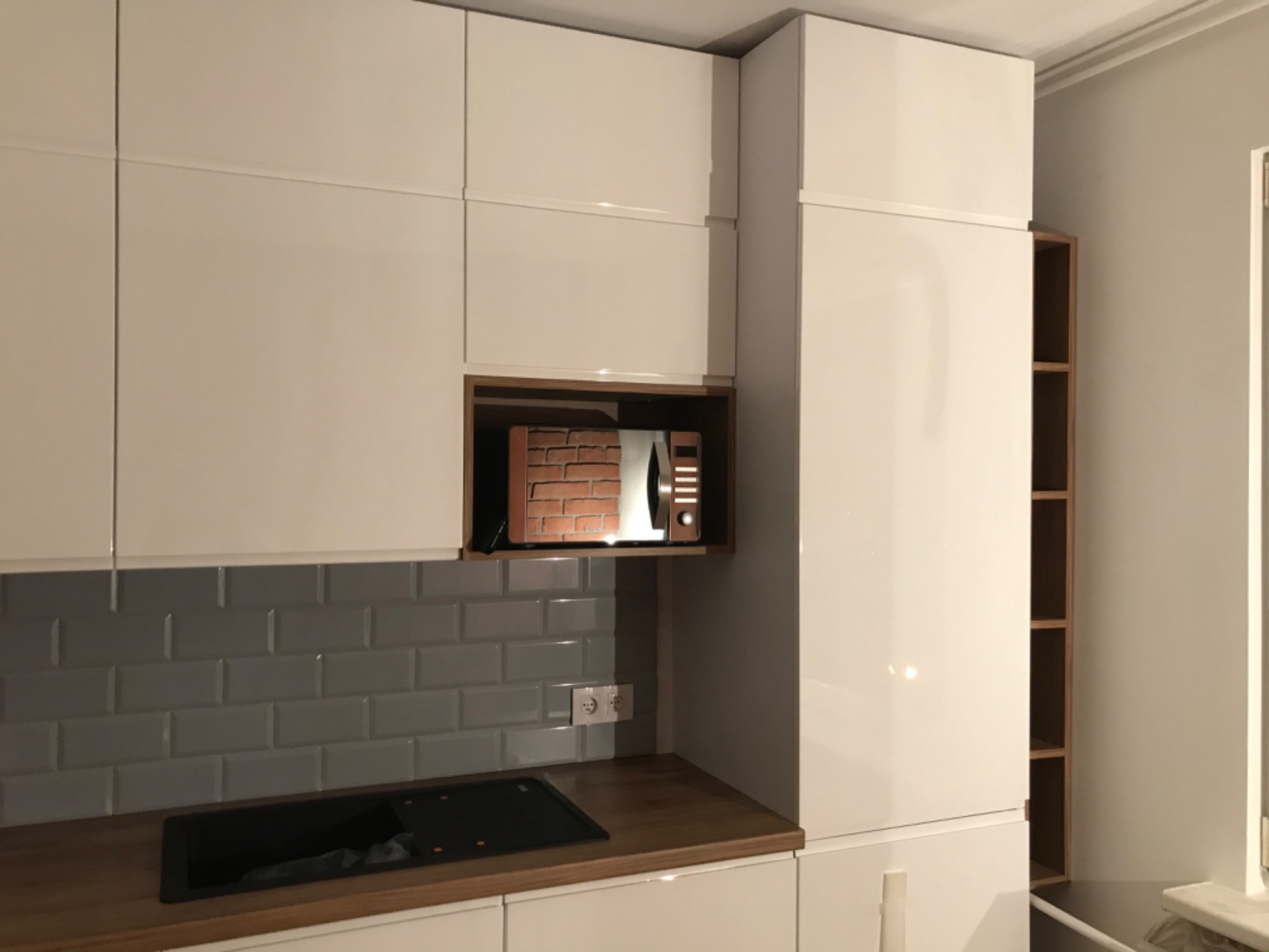 Nowoczesna biała kuchnia z połyskiem, szare kafelki nad blatem, drewniany blat, płyta indukcyjna i zabudowana mikrofalówka w drewnianej wnęce.