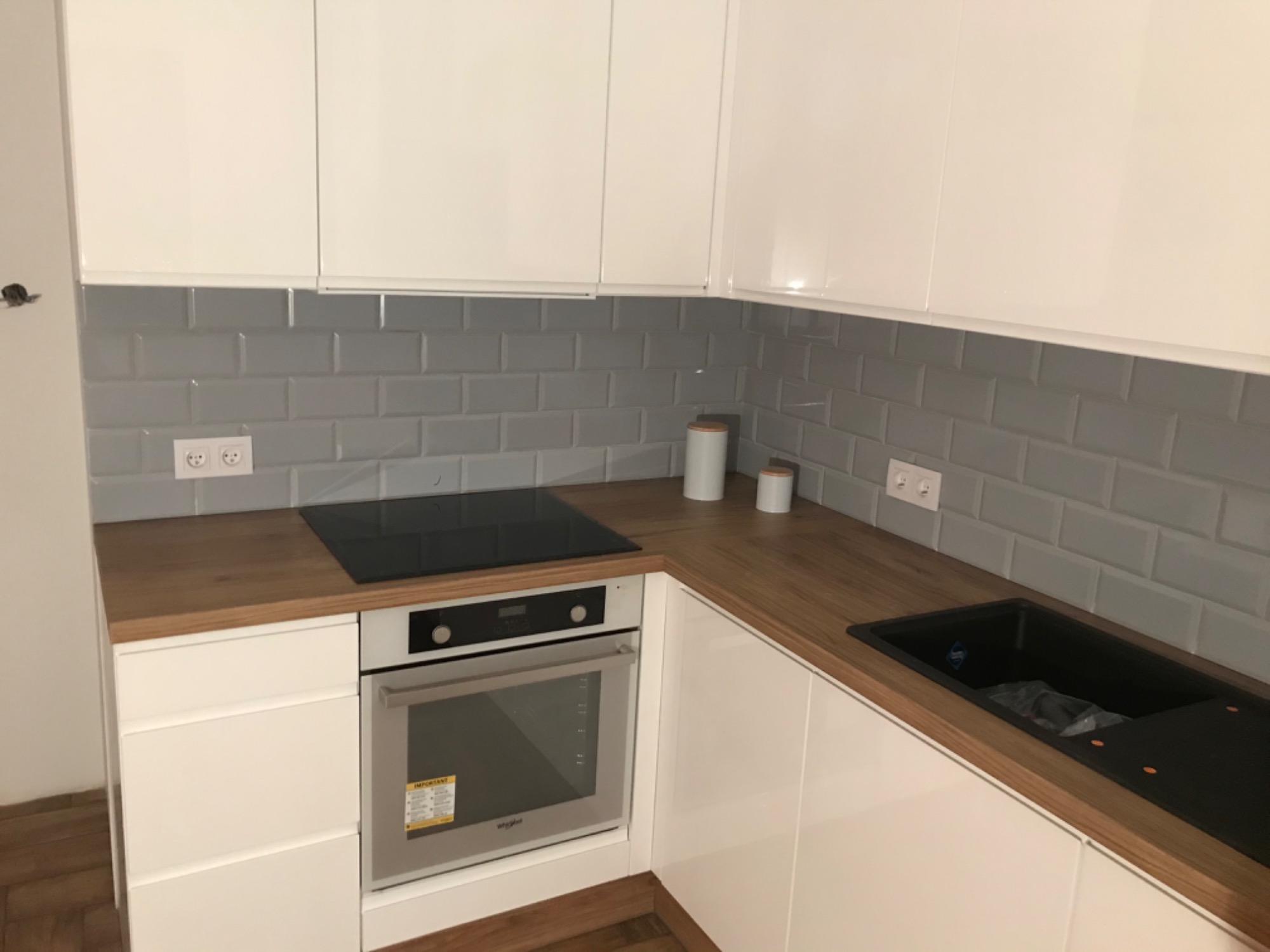 Nowoczesna kuchnia z białymi, lakierowanymi frontami, drewnianym blatem, szarą glazurą typu metro i czarnym zlewem kompozytowym.