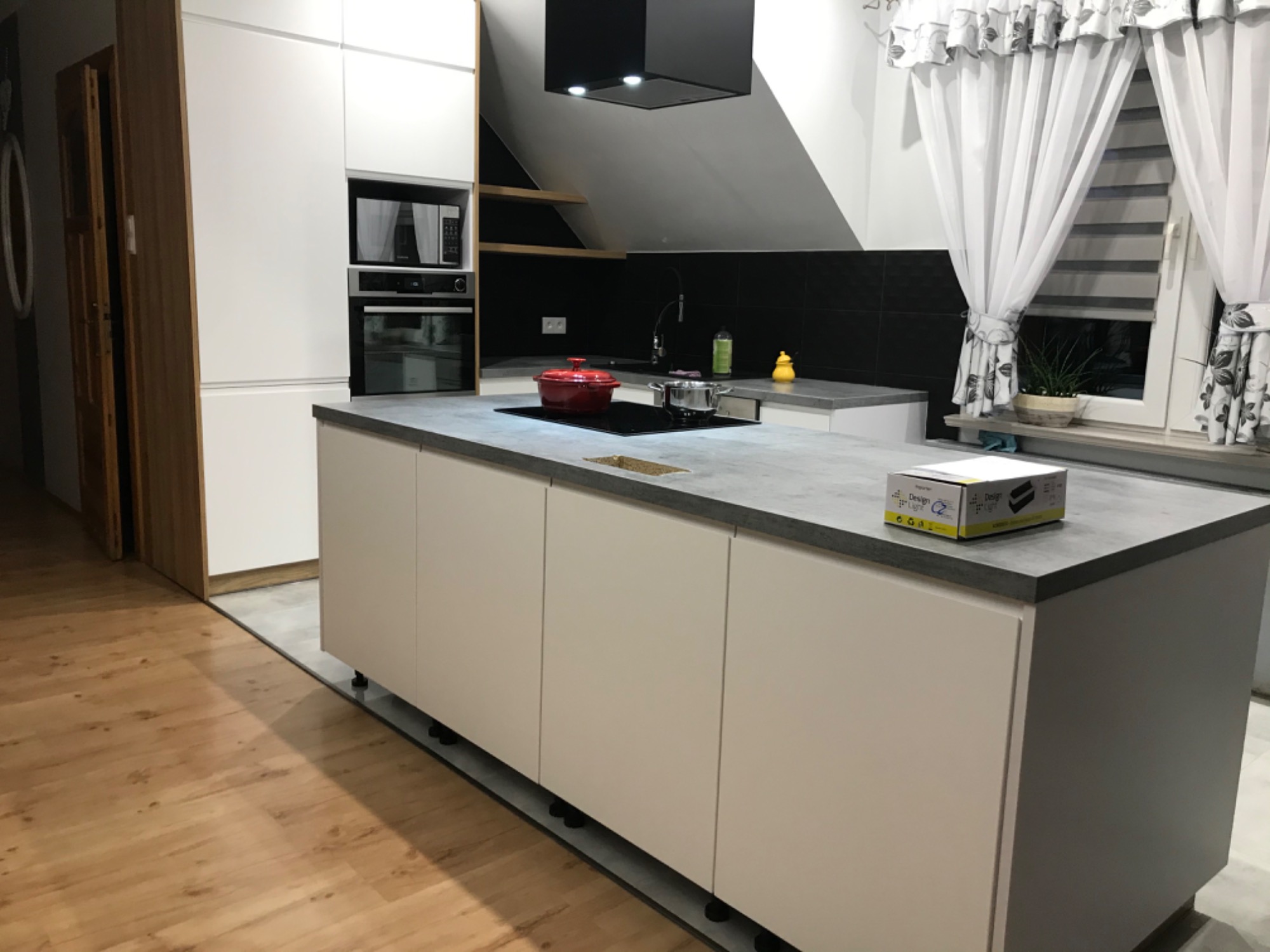 Nowoczesna kuchnia z białymi frontami szafek i szarym blatem, na którym stoi czerwony garnek i srebrny rondel. Widoczny czarny okap, czarna ściana nad blatem oraz okno z firankami w kwiaty.